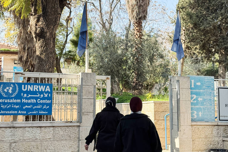 A imagem mostra a entrada do Centro de Saúde da UNRWA em Jerusalém. Duas pessoas estão caminhando em direção ao portão， que é cercado por árvores e bandeiras azuis. O letreiro à esquerda exibe o nome do centro em inglês， árabe e francês.