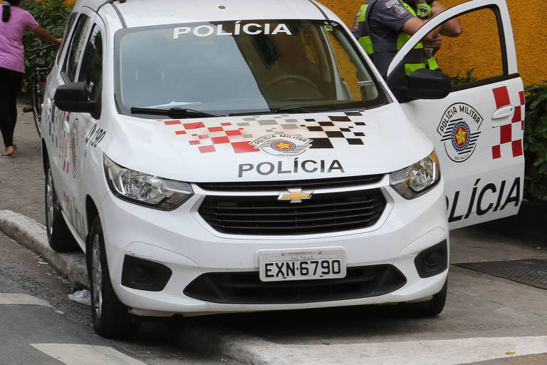 Imagem de uma van da Polícia Militar estacionada em uma rua. O veículo é branco com detalhes em vermelho e preto， e possui a palavra 039;POLÍCIA039; em destaque na parte superior. É possível ver uma pessoa caminhando e um policial próximo à porta do veículo.