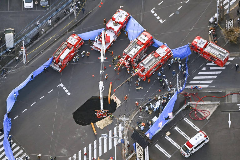 A imagem mostra um cruzamento urbano onde várias viaturas de bombeiros estão posicionadas em torno de um buraco no chão. Há uma faixa azul delimitando a área do acidente， e algumas pessoas estão visíveis na cena. O ambiente é urbano， com faixas de pedestres e veículos estacionados ao fundo