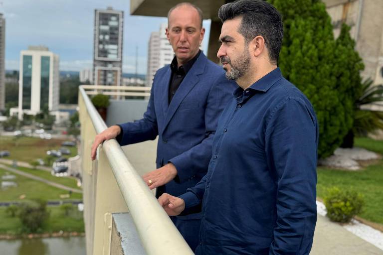 A imagem mostra dois homens em um terraço. Eles estão de pé ao lado de uma grade， olhando para a paisagem. O homem à esquerda está usando uma jaqueta escura e o homem à direita está vestido com uma camisa azul. Ao fundo， há edifícios modernos e uma área verde.