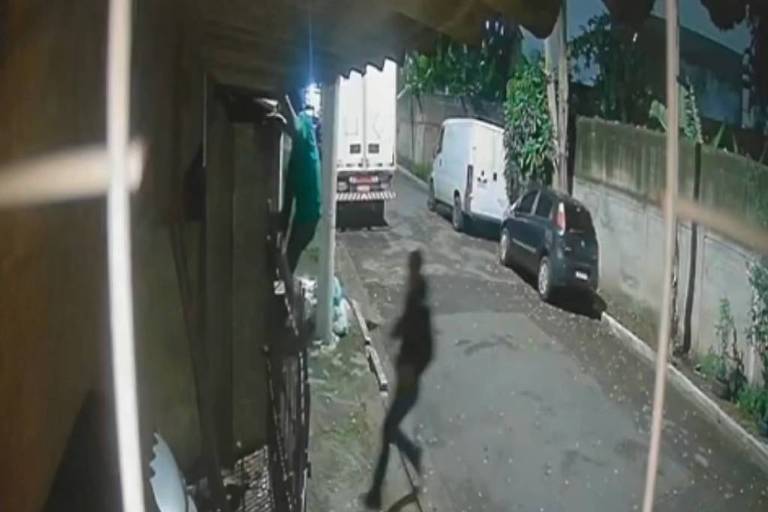 A imagem mostra uma cena noturna capturada por uma câmera de segurança. À esquerda， uma pessoa vestindo uma camisa verde está subindo em uma estrutura， enquanto um caminhão branco está estacionado ao fundo. Há também uma van branca e um carro preto estacionados na rua. A iluminação é baixa， e a sombra de uma pessoa pode ser vista caminhando na calçada.