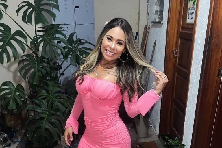 A imagem mostra uma mulher em pé em um ambiente interno， usando um vestido rosa justo e de mangas longas. Ela está sorrindo e segurando o cabelo， com plantas ao fundo e uma porta de madeira ao lado.