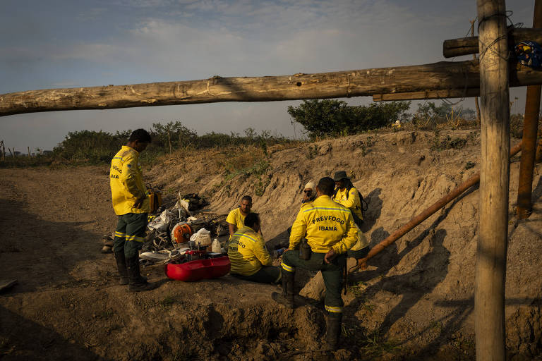 A imagem mostra uma equipe de resgate vestindo uniformes amarelos， reunida em um local ao ar livre. Eles estão sentados e em pé ao redor de equipamentos de resgate， com um fundo de terreno arenoso e vegetação escassa. A luz do sol ilumina a cena， criando um ambiente de trabalho ativo.