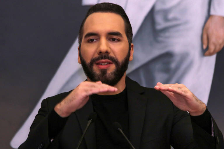 Imagem mostra Nayib Bukele， presidente de El Salvador， um homem com barba e cabelo curto， vestindo uma jaqueta preta， e que está em pé em frente a um fundo com uma imagem de uma pessoa vestindo roupas brancas. Ele está gesticulando com as mãos， fazendo um movimento horizontal， enquanto fala em um microfone.