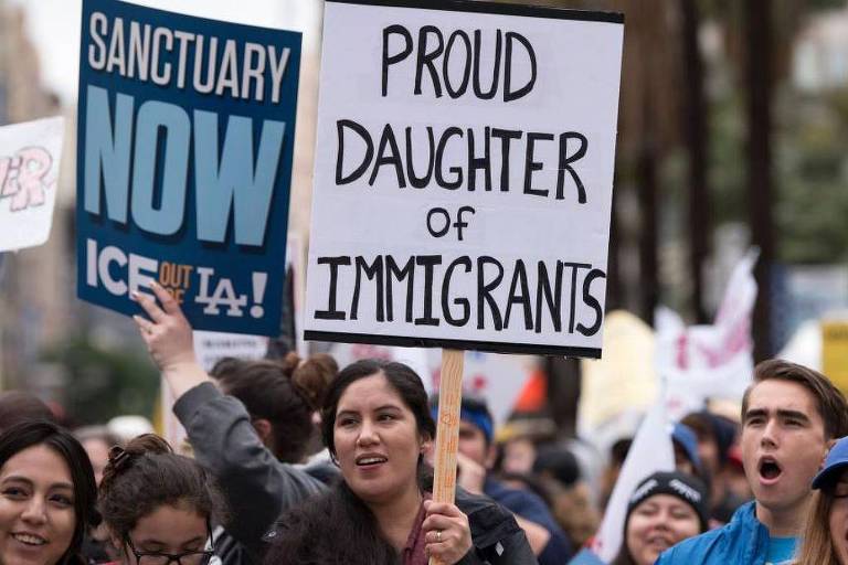 A imagem mostra um grupo de pessoas participando de um protesto. No centro， uma mulher segura um cartaz que diz 039;PROUD DAUGHTER OF IMMIGRANTS039;. Ao fundo， há outros cartazes， incluindo um que diz 039;SANCTUARY NOW039; e outro com a frase 039;ICE OUT LA!039;. As pessoas parecem estar unidas em uma causa， com expressões de determinação.