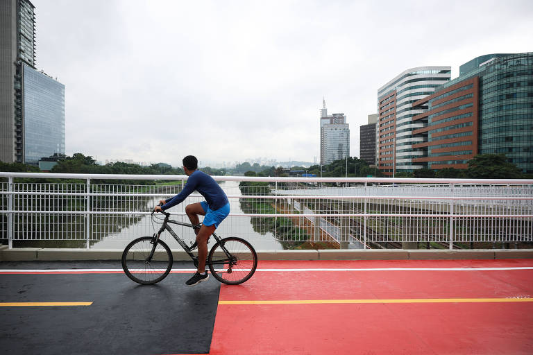 Um ciclista está parado em uma bicicleta em uma ponte, olhando para a paisagem urbana. O ciclista usa uma camiseta azul e shorts azuis. Ao fundo, há edifícios modernos e um céu nublado. A ponte tem uma faixa de ciclistas em vermelho e preto.
