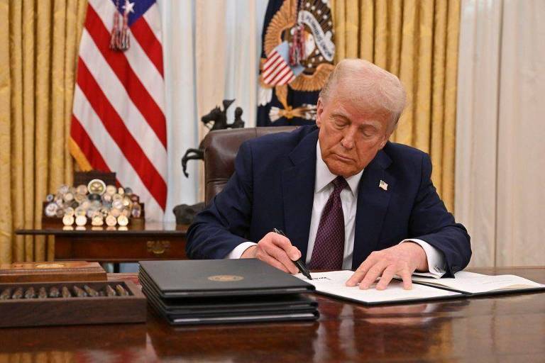 Um homem sentado em uma mesa， escrevendo em um documento. Ele está usando um terno escuro e uma gravata. Ao fundo， há uma bandeira dos Estados Unidos e um emblema presidencial. Na mesa， há vários documentos empilhados e um objeto decorativo com medalhas.