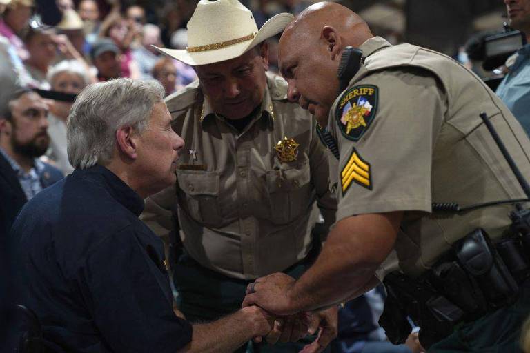 A imagem mostra três homens em um ambiente público. Um deles， à esquerda， está sentado e parece estar conversando com os outros dois， que estão em pé. O homem à direita é um policial com um distintivo e um cinto de utilidades， enquanto o homem no meio usa um chapéu de cowboy e uma camisa de uniforme. O fundo é desfocado， mas há uma multidão visível.