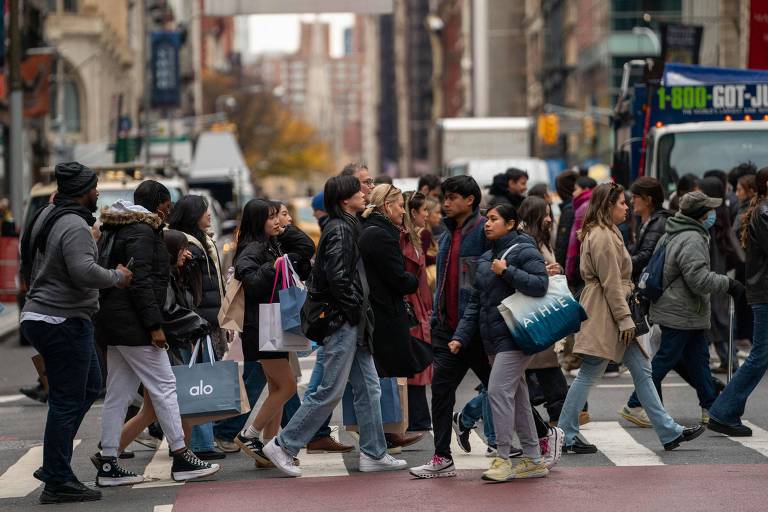 A imagem mostra um grupo de pessoas atravessando uma rua em um cruzamento urbano. As pessoas estão vestidas de maneira variada， algumas com sacolas de compras. Ao fundo， é possível ver edifícios e um ônibus estacionado. O clima parece ser de um dia nublado.