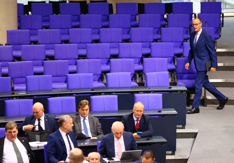 A imagem mostra um ambiente de uma sessão legislativa， com várias cadeiras roxas vazias ao fundo. No primeiro plano， há um grupo de homens sentados em mesas， alguns usando ternos escuros e gravatas. Um homem em um terno azul está caminha em outra direção.