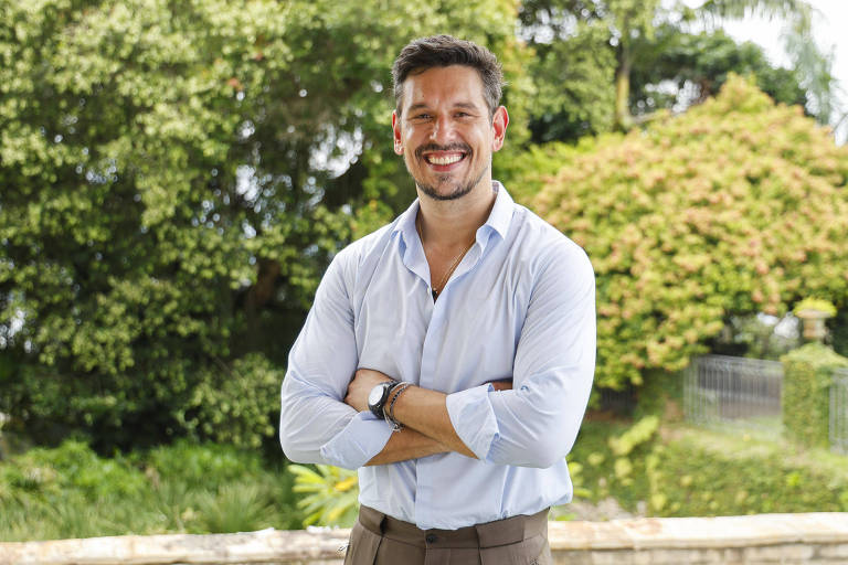 Um homem está posando com os braços cruzados e sorrindo. Ele usa uma camisa de botão azul clara e calças marrons. Ao fundo, há uma vegetação densa e verde, com árvores e arbustos, criando um ambiente natural e agradável.
