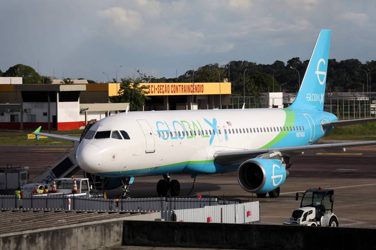 Um avião da companhia aérea com uma pintura em tons de azul e verde está estacionado em um aeroporto. Ao fundo， é possível ver um terminal e uma pista de pouso. O céu está parcialmente nublado