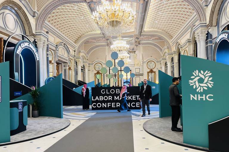 A imagem mostra o interior de um grande salão decorado para a Conferência Global do Mercado de Trabalho. Há painéis verdes com textos e logotipos， um tapete cinza no chão e um lustre elegante no teto. Algumas pessoas estão presentes no espaço， que possui uma arquitetura elaborada.