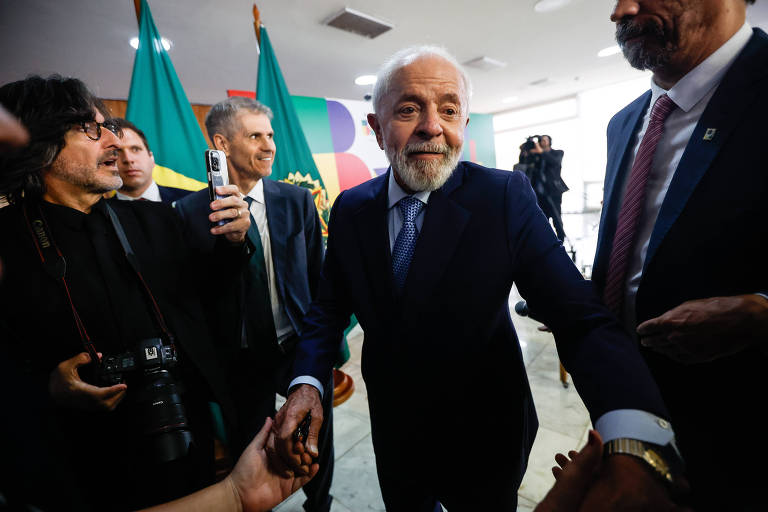 A imagem mostra um homem idoso de barba branca， vestindo um terno escuro e gravata， sorrindo enquanto cumprimenta alguém. Ele está cercado por várias pessoas， algumas segurando câmeras e celulares. Ao fundo， há bandeiras do Brasil， sugerindo um evento oficial ou político. O ambiente é bem iluminado e parece movimentado.