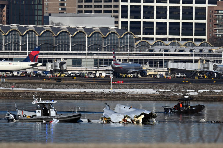 A imagem mostra uma cena de um acidente aéreo em uma área urbana， com uma aeronave parcialmente submersa na água. Vários barcos de resgate estão próximos à aeronave， enquanto ao fundo se vê um terminal de aeroporto e aviões estacionados. O ambiente é urbano， com edifícios altos ao fundo.