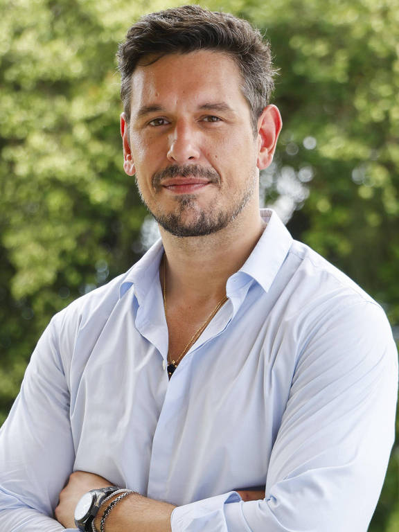 Um homem está posando com os braços cruzados e sorrindo. Ele usa uma camisa de botão azul clara e calças marrons. Ao fundo, há uma vegetação densa e verde, com árvores e arbustos, criando um ambiente natural e agradável.