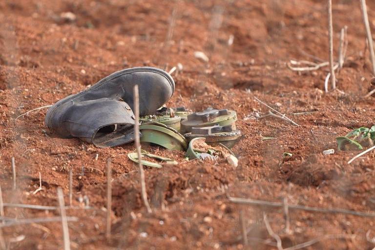 A imagem mostra um par de sapatos pretos， um deles virado para cima， e alguns objetos metálicos， incluindo uma parte de um dispositivo， em um solo de terra avermelhada. O ambiente parece seco e árido， com algumas pequenas plantas secas ao redor.