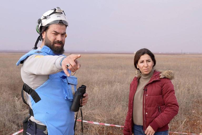 A imagem mostra um homem e uma mulher em um campo aberto. O homem， com uma barba e usando um colete azul e um capacete， está apontando para algo à sua frente. Ele segura um dispositivo de comunicação. A mulher， com cabelo escuro e vestindo uma jaqueta vermelha， observa atentamente. O fundo é uma vasta área de vegetação seca sob um céu nublado.