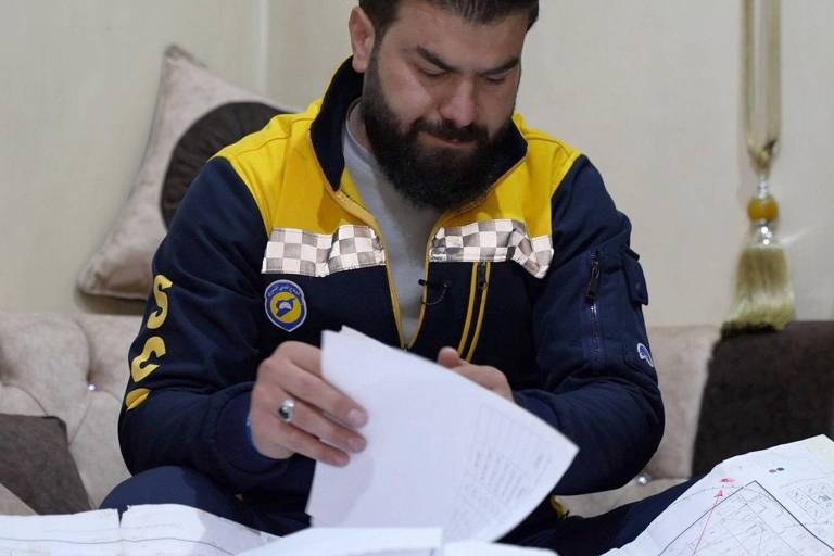 Um homem com barba e cabelo curto está sentado em um sofá， analisando papéis e documentos. Ele veste uma jaqueta azul com detalhes amarelos e um emblema no peito. O ambiente é interno， com uma decoração simples ao fundo.