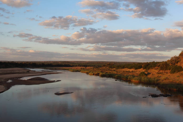 A imagem mostra um rio calmo refletindo o céu ao entardecer. O céu está parcialmente nublado， com nuvens em tons de azul e laranja. À margem do rio， há vegetação e áreas de areia exposta. O ambiente transmite uma sensação de tranquilidade e beleza natural.
