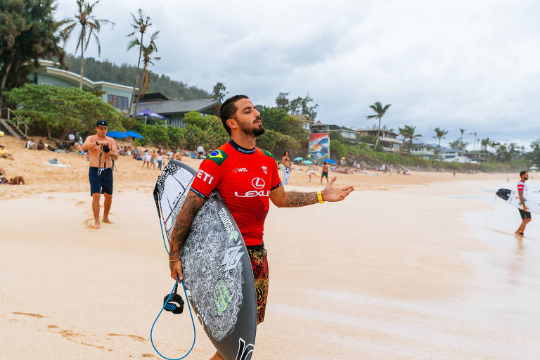 Um surfista em pé na areia da praia， vestindo uma camiseta vermelha. Ele segura uma prancha de surfe e parece estar se preparando para entrar na água. Ao fundo， há outras pessoas na praia e vegetação ao redor. O céu está nublado.