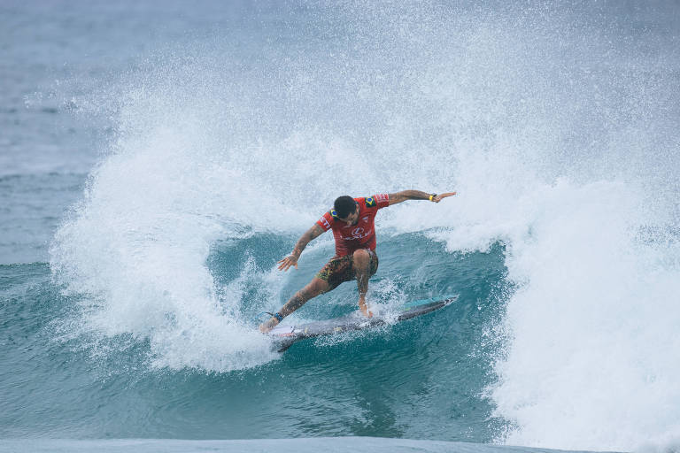 Um surfista está pegando uma onda， realizando uma manobra sobre a prancha. Ele usa uma camiseta vermelha e está cercado por água espumante. O céu está nublado e as ondas são grandes.