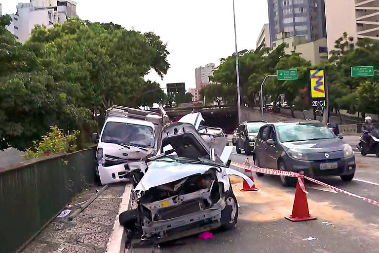 A imagem mostra um acidente de trânsito em uma via urbana. Um carro está danificado e parcialmente fora da pista， com a parte frontal amassada. Há um veículo branco ao fundo， também danificado， e outros carros parados na via. Cone de sinalização estão posicionados ao redor da área do acidente， e há uma vegetação ao lado da via.