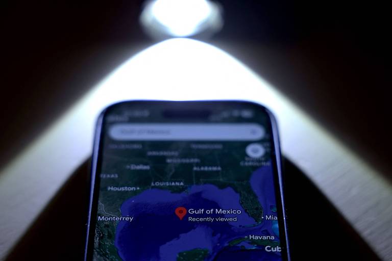 A imagem mostra um smartphone com a tela voltada para cima， exibindo um mapa. A localização marcada é 039;Gulf of Mexico039;. Há uma fonte de luz iluminando a parte superior do telefone， criando um efeito de destaque. O fundo é escuro.
