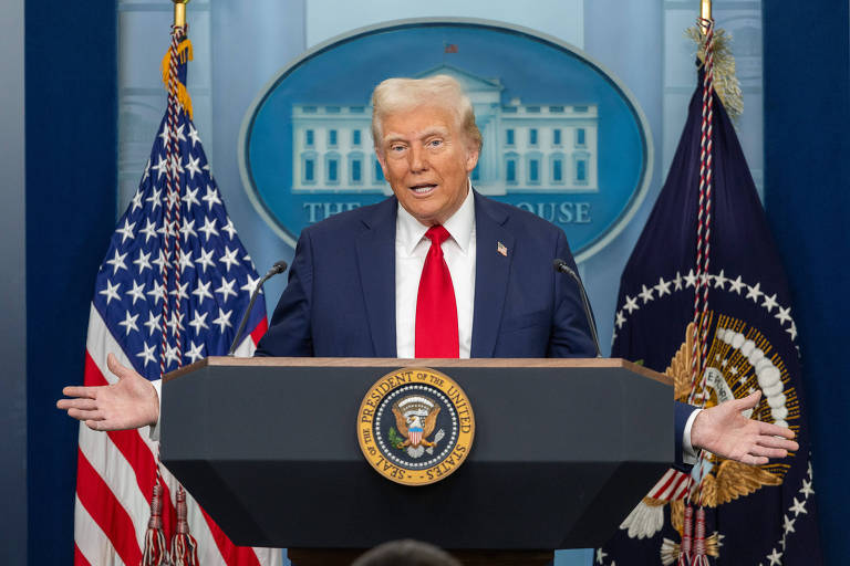 Um homem em um palanque de conferência， com bandeiras dos Estados Unidos ao fundo. Ele está usando um terno escuro e uma gravata vermelha， gesticulando com as mãos enquanto fala. Ao fundo， há um emblema presidencial e uma parede azul.