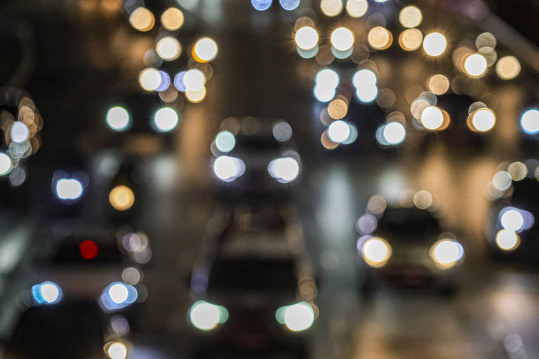 A imagem mostra uma cena de tráfego noturno， com muitos carros visíveis. As luzes dos faróis e lanternas estão desfocadas， criando um efeito bokeh， onde as luzes aparecem como círculos brilhantes. A estrada parece movimentada， mas a falta de nitidez impede a identificação clara dos veículos.