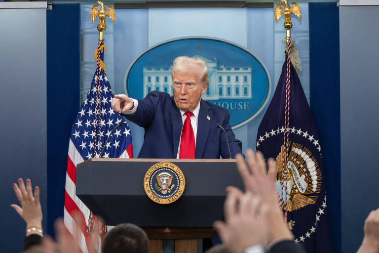 Um homem em um palanque， apontando com a mão， enquanto várias mãos levantadas estão visíveis na frente. Ao fundo， bandeiras dos Estados Unidos e um emblema presidencial. O ambiente parece ser uma sala de imprensa da Casa Branca.
