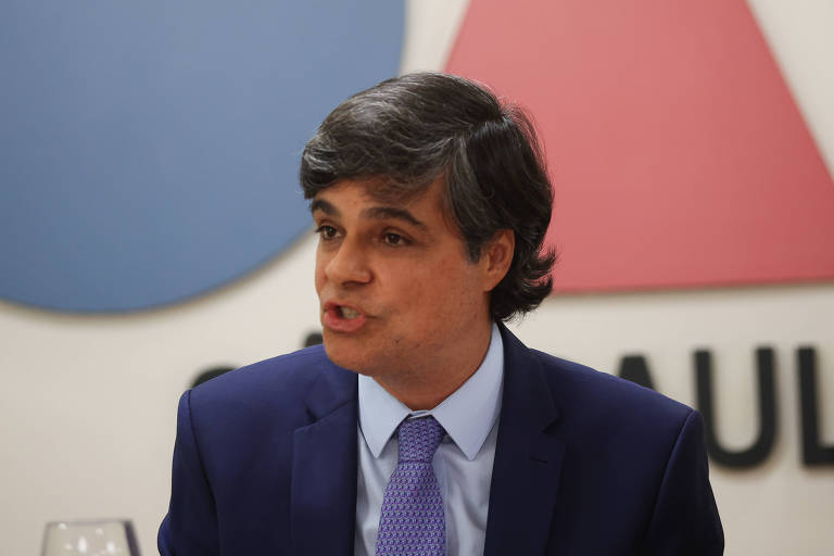 Um homem com cabelo castanho e liso， vestido com um terno escuro e uma gravata lilás， está falando em um evento. Ao fundo， há um logotipo em cores azul e vermelho， com a palavra 039;SÃO PAULO039; visível.
