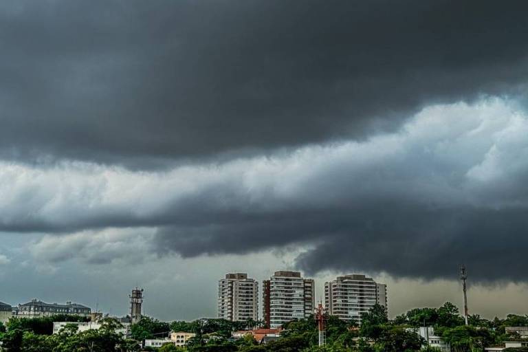 A imagem mostra um céu nublado， com nuvens escuras e volumosas. Ao fundo， há vários edifícios de vários andares， e a paisagem é urbana. A luz é difusa， sugerindo que pode estar prestes a chover.