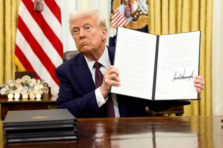 Um homem está sentado em uma mesa， segurando um documento em uma mão e olhando para a câmera. Ao fundo， há uma bandeira dos Estados Unidos e outros itens decorativos. O homem tem cabelo loiro e está vestido com um terno escuro. Na mesa， há vários documentos empilhados.
