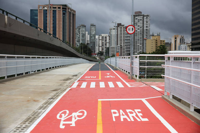 A imagem mostra uma ciclovia pintada de vermelho， com sinalização de bicicleta e a palavra 039;PARA039; em branco. Ao fundo， há prédios altos e um céu nublado， indicando um ambiente urbano. A ciclovia é ampla e está localizada em uma ponte ou via elevada.