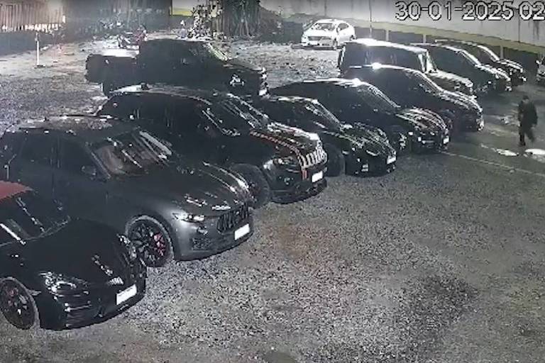 A imagem mostra um estacionamento à noite com vários carros， predominantemente pretos. Os veículos estão alinhados em uma superfície de cascalho， e há uma iluminação suave ao fundo. Um carro vermelho se destaca entre os outros. A cena é tranquila e bem iluminada.