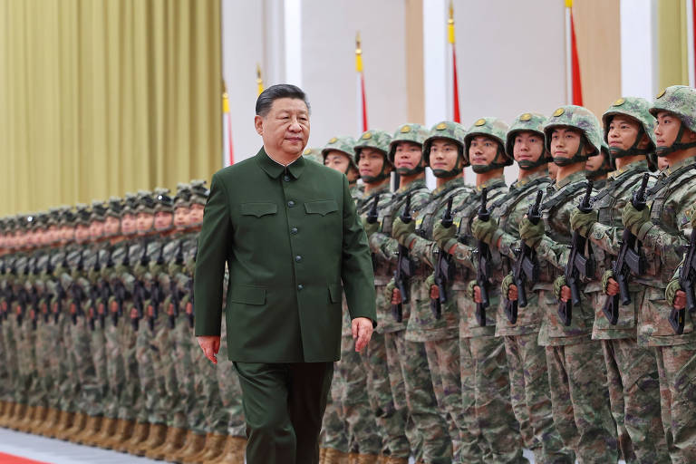 A imagem mostra um líder militar caminhando em frente a uma fileira de soldados em trajes de combate. Os soldados estão alinhados em formação， com armas e uniformes camuflados. O ambiente é formal， com bandeiras ao fundo e uma decoração simples.