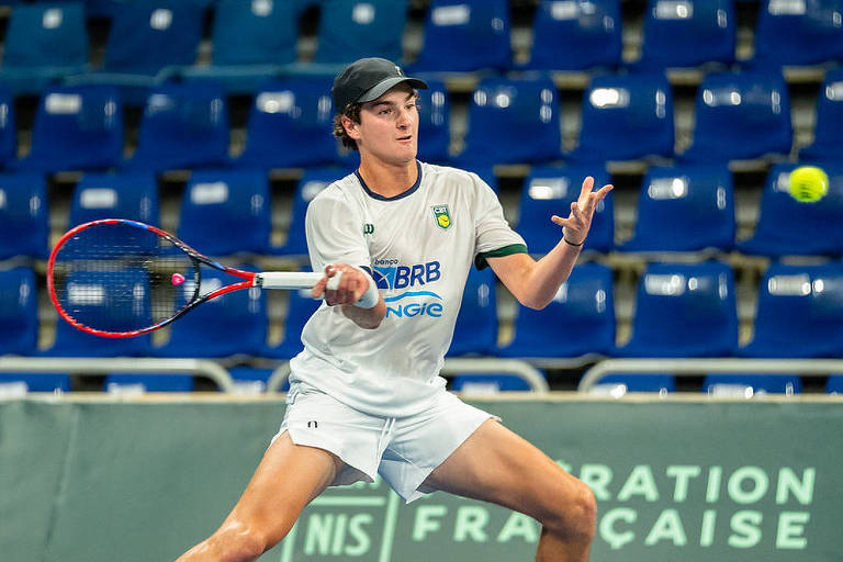 Um jovem jogador de tênis está em ação， prestes a rebater uma bola. Ele usa uma camiseta branca com detalhes em verde e azul， e um boné preto. O fundo mostra uma arquibancada vazia com cadeiras azuis. O jogador está concentrado e em movimento， segurando uma raquete de tênis.