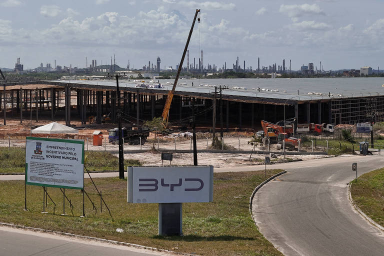 A imagem mostra um canteiro de obras da fábrica da BYD， com estruturas de construção em andamento. Há guindastes e materiais de construção visíveis. No primeiro plano， há um grande painel com o logotipo da BYD e informações sobre o projeto. Ao fundo， é possível ver uma área industrial com várias instalações.