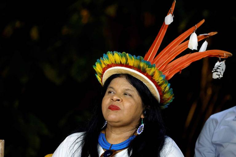 Sonia, mulher indígena, aparece do ombro para cima. Ela usa cocar colorido e blusa branca com colar azul. Fundo da foto é escuro.