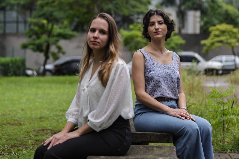 A imagem mostra duas mulheres sentadas em um banco em um parque. A mulher à esquerda tem cabelo longo e liso， vestindo uma blusa branca e calças escuras. A mulher à direita tem cabelo curto e ondulado， usando uma blusa cinza e calças jeans claras. Ao fundo， há árvores e carros estacionados