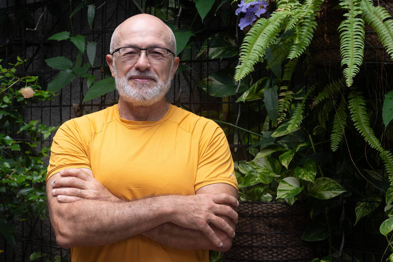 Um homem de cabelos grisalhos e barba， usando uma camiseta laranja， está posando com os braços cruzados em frente a um fundo de plantas verdes e flores. Ele usa óculos e tem uma expressão serena.