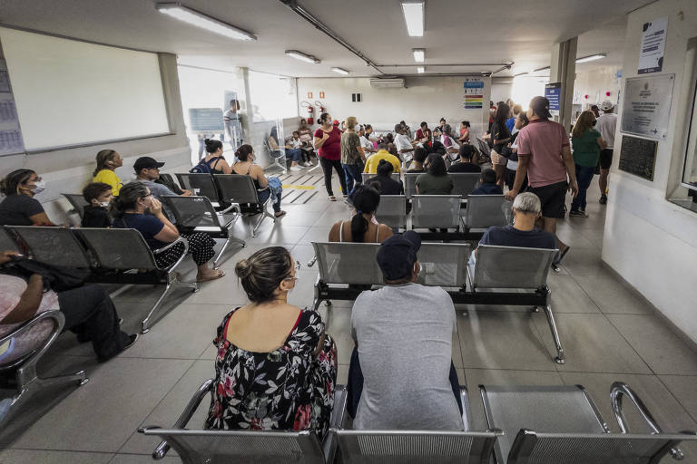 A imagem mostra uma sala de espera em um hospital， com várias pessoas sentadas em cadeiras de metal. Algumas pessoas estão usando máscaras. No fundo， há um grupo de pessoas em pé， possivelmente aguardando atendimento. O ambiente é bem iluminado e possui paredes brancas.