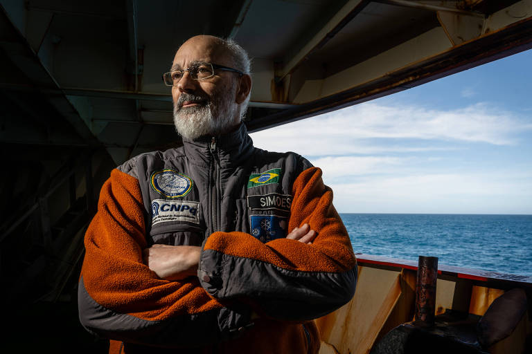 Um homem de cabelos grisalhos e barba， usando óculos， está em um navio. Ele veste um casaco cinza com detalhes em laranja e possui emblemas em seu traje. O homem está com os braços cruzados e olhando para o horizonte， com o mar visível ao fundo e um céu parcialmente nublado.