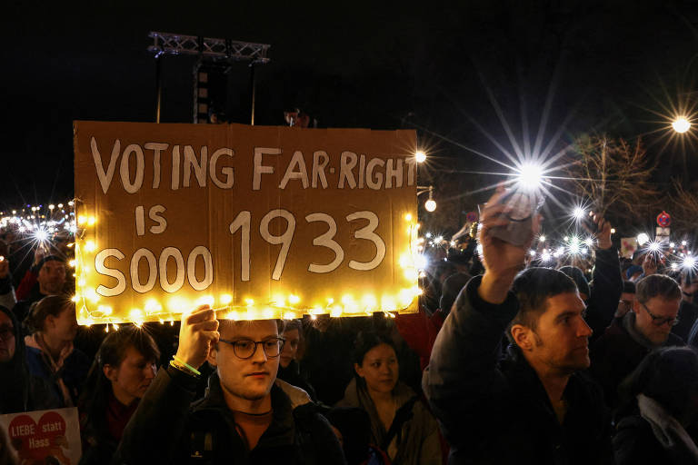 A imagem mostra um protesto à noite， onde várias pessoas seguram placas e celulares com luzes acesas. Em destaque， uma pessoa segura uma placa iluminada que diz 039;VOTING FAR RIGHT IS sooo 1933039;. O ambiente é de agitação e as luzes criam um efeito visual marcante.