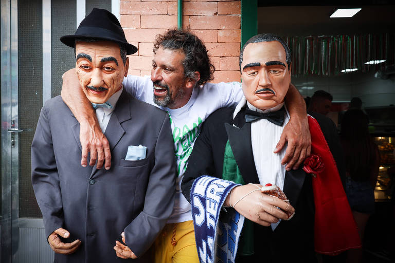 Um homem sorridente está posando entre dois bonecos grandes, que representam figuras masculinas vestidas de forma formal. O homem está usando uma camiseta branca e calças amarelas, enquanto os bonecos estão vestidos com trajes de gala, um com um chapéu e o outro com uma capa vermelha. O fundo é uma parede de tijolos e há luzes artificiais visíveis.