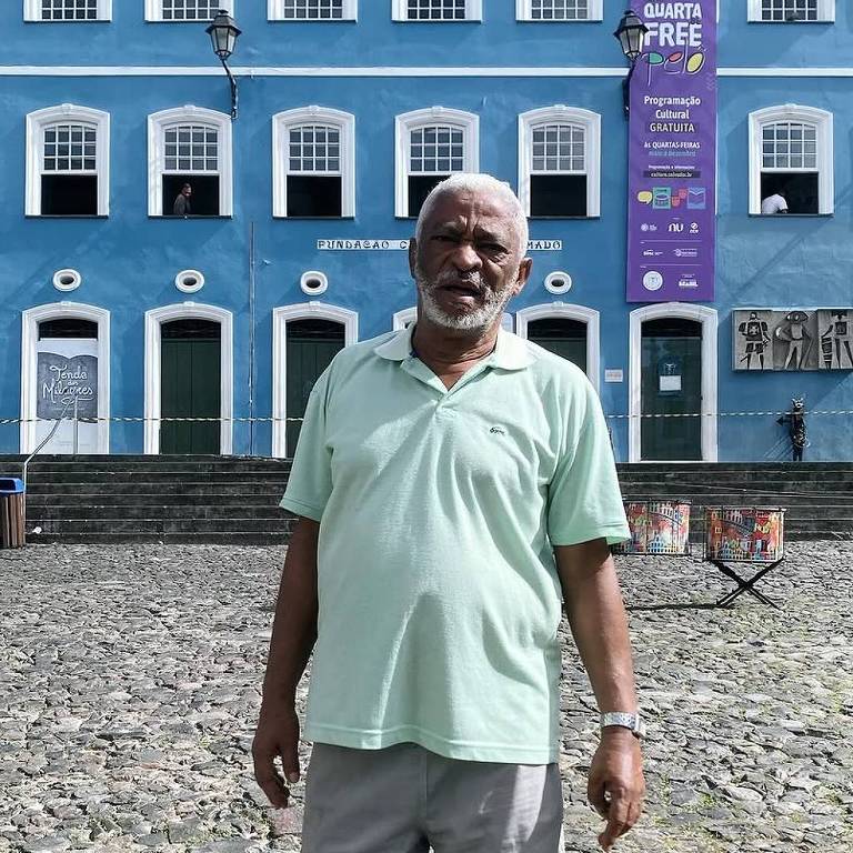 Um homem idoso está posando em uma praça， vestindo uma camiseta verde clara e shorts. Ele está descalço e sorri para a câmera. Ao fundo， há um edifício de cor azul com janelas brancas e uma faixa colorida pendurada. O chão é de pedras irregulares.