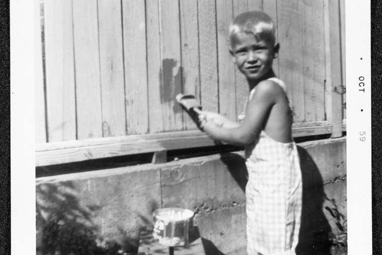 A imagem mostra uma criança com cabelo loiro e curto, usando um macacão xadrez, pintando uma cerca de madeira. A criança está segurando um pincel e sorrindo, enquanto aplica tinta em uma parte da cerca. Ao lado, há um balde de tinta. O fundo é composto por uma parede de concreto e algumas plantas.