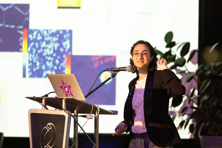 Uma mulher está em pé em um palco， falando em um microfone. Ela usa um blazer preto e sorri enquanto gesticula com a mão. À sua frente， há um laptop sobre um suporte e um fundo com várias imagens coloridas e gráficos. Plantas podem ser vistas ao fundo.