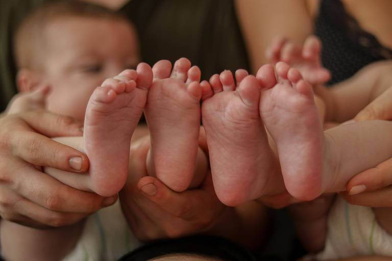 A imagem mostra os pés de dois bebês sendo segurados por mãos adultas. Os pés estão em primeiro plano， com a pele clara e dedos pequenos. Ao fundo， é possível ver parcialmente os rostos dos bebês， mas estão desfocados.